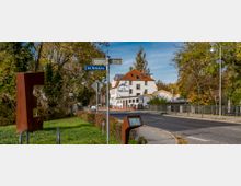 Straßenszene mit einem Wegweiser „Am Nottehafen“, einem modernen, rostfarbenen Kunstobjekt aus Metall und einem weißen Gebäude mit rotem Dach, das ein Hotel oder Restaurant beherbergt. Im Hintergrund Bäume und ein Geländer entlang des Notte-Kanals.