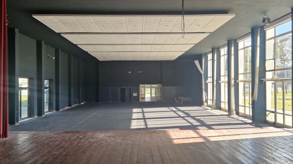 Ein leerer Veranstaltungsraum mit einer erhöhten Bühne aus Holz im Vordergrund, hohen Fenstern an der rechten Seite, die viel Tageslicht hereinlassen, sowie einer dunklen Wand am Ende des Raumes mit einer Doppeltür in der Mitte. Die Decke ist mit rechteckigen Akustikplatten verkleidet.