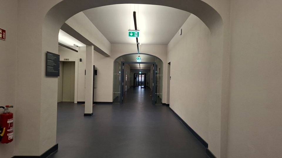 Langer, weiß gestrichener Innenflur in einem öffentlichen Gebäude mit Rundbögen, dunkelgrauem Boden und Deckenleuchten. In der Mitte führen gläserne Doppeltüren weiter in den Korridor, darüber sind grüne Notausgangsschilder angebracht.