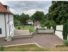 Ansicht eines Hofs mit einer weißen Villa links im Vordergrund, roten Ziegeldächern und blauen Fensterläden. Im Hintergrund befinden sich ein kleines Häuschen mit rotem Dach, eine Rasenfläche, hohe Bäume und ein angrenzender Zaun, der das Grundstück umgibt.