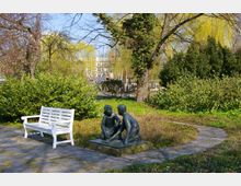 Eine Parkszene mit einer weißen Holzbank und einer Statue zweier kniender Kinder auf einem Sockel. Im Hintergrund sind dichtes Gebüsch, hohe Bäume und ein Spielplatz mit Holzstrukturen zu sehen.