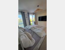 Helles Schlafzimmer mit großem Bett und weißer Bettwäsche, grauen Vorhängen und bodentiefen Fenstern, von denen eines als Balkontür geöffnet ist. An der Wand hängt ein Flachbildfernseher, an der Decke eine geflochtene Pendelleuchte; draußen sind Grünflächen und Nachbargebäude sichtbar.