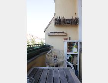 Small apartment balcony with a wooden table and white metal chair beside a railing lined with empty planter boxes. A pale stucco wall has rustic wooden shelves and a small white storage cabinet holding pots and gardening supplies, with neighboring rooftops visible in the background.