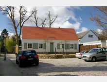 Ein kleines, weißes Gebäude mit rotem Satteldach, grünen Fensterläden und einer Eingangstür, umgeben von kahlen Bäumen. Im Vordergrund liegt ein gepflasterter Platz, auf dem mehrere Autos geparkt sind.