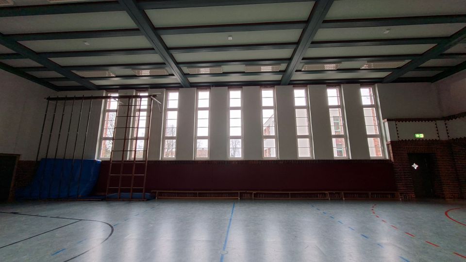 Innenansicht einer Sporthalle mit hohem Rasterdecken-Tragwerk, großer Fensterfront aus schmalen, hohen Fenstern und markierten Hallenbodenlinien. An der Wand stehen Kletterstangen und eine Sprossenwand mit blauen Turnmatten; rechts ist ein Ausgang in einer Ziegelwand.