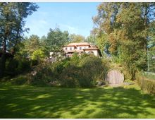Zweistöckiges Haus mit hellem, schrägem Dach, umgeben von Bäumen und dichter Vegetation. Im Vordergrund befindet sich eine Wiese, rechts ein halbrundes Holztor, das teilweise in die Erde eingebaut ist.