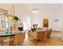 Helles Wohnzimmer mit Holzboden, einer gemütlichen Sitzecke mit beigen Sesseln und Sofa, einem runden Esstisch mit Stühlen sowie einem dekorativen Kaminsims aus weißem Marmor. Im Raum stehen moderne Hängelampen und Pflanzen, im Hintergrund ist eine geöffnete Tür zur angrenzenden Küche zu sehen.