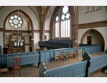 Innenraum einer Kirche mit Backsteinbögen und hohen Fenstern mit farbigen Glasfassaden. Im Vordergrund hölzerne Kirchenbänke und Stühle, im Hintergrund ein Altar mit Kunstwerken sowie Galerie-Bereiche an den Seiten.