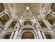 Innenansicht eines historischen Gebäudes mit prächtiger barocker Architektur, fein verzierten Säulen und Bögen. Die Decke zeigt ein kunstvolles Gewölbemuster, während Statuen und aufwendige Ornamentik die Wände schmücken.