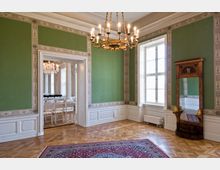 Ein elegant gestalteter Raum mit grünen Wänden, dekorativen weißen Bordüren und einem verzierten Kronleuchter. Der Raum hat ein großes Fenster, einen Holzboden mit einem Teppich und einen stilvollen Wandspiegel; durch eine geöffnete Tür ist ein angrenzender Raum mit Stühlen und einem weiteren Kronleuchter sichtbar.