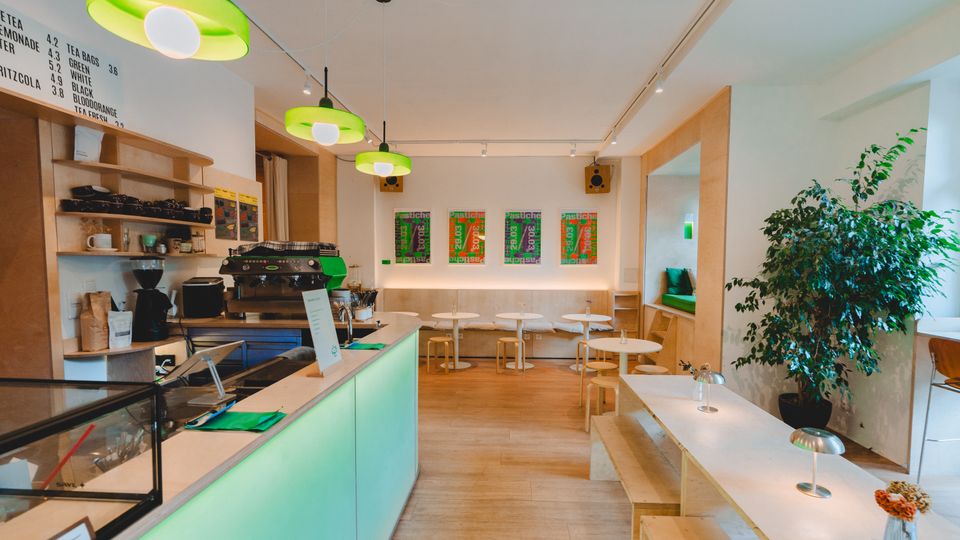 Modern café interior with a mint-green service counter, open wood shelving, and a seating area of small round tables and long benches on light wood floors. Green pendant lights hang from the ceiling, colorful posters line the back wall, and a large potted plant sits near a windowed nook.