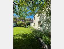 Innenhofgarten mit großer Rasenfläche, umgeben von Bäumen und Sträuchern, mit einer Terrasse und Sonnenschirm im Hintergrund. Rechts steht ein hell verputztes Wohngebäude mit mehreren Fenstern, im Hintergrund ist ein mehrstöckiges Backsteingebäude zu sehen.