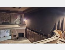 Verlassener Theater- oder Kinosaal mit leerer Bühne, einem Balkon mit Geländer und stark abgenutzten, teilweise abgeplatzten Wänden. Rechts dominiert eine große dunkle Wand mit freiliegenden Kabeln und Baustellenleuchte; der Boden ist staubig und stellenweise beschädigt.