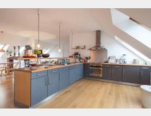 Moderne Küche in einem Dachgeschoss mit blauen Unterschränken, Arbeitsplatten aus Holz und schrägen Dachfenstern. Im Raum befinden sich hängende Glühbirnen, eine Dunstabzugshaube sowie diverse Küchengeräte und Dekorationen.