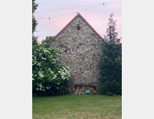 Steinwand eines kleinen Gebäudes mit einem spitz zulaufenden Ziegeldach, flankiert von einem blühenden Strauch auf der linken Seite und einem Baum auf der rechten Seite; davor gestapeltes Brennholz und eine Pflanze in einem blauen Topf. Im Hintergrund ein Himmel mit rosa Licht und eine Lichterkette.