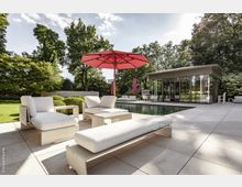 Moderne Terrasse mit hellen Lounge-Möbeln und großem roten Sonnenschirm neben einem rechteckigen Pool. Im Hintergrund ein Gebäude mit Glasfronten, umgeben von grünen Bäumen und gepflegter Gartenlandschaft.