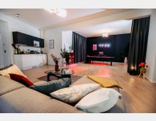 Offener Wohnraum mit moderner Ausstattung: Im Vordergrund ein graues Sofa mit bunten Kissen und einem Glastisch, dahinter eine offene Küche mit weißen Schränken und schwarzer Front. Im Hintergrund ein Bereich mit schwarzer Wand, einem DJ-Pult mit Lichteffekten und dekorativen Pflanzen.