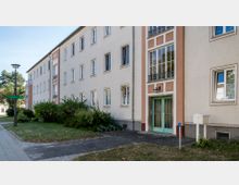 Mehrstöckiges Wohngebäude im Stil der Nachkriegsarchitektur mit cremefarbener Fassade, rosa Akzenten und einem Eingangsbereich mit grünen Glastüren. Davor befindet sich eine kleine Grünfläche mit Büschen und einem Gehweg.