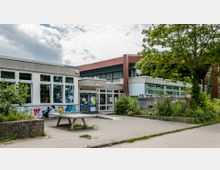 Flaches Gebäude mit großen Fenstern, bemalten Wänden und Graffiti an der Vorderseite, umgeben von Bäumen und Pflanzen. Davor ein kleiner gepflasterter Platz mit einer Holzbank und einem Tisch.