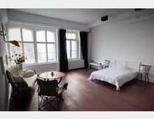 Ein schlicht eingerichtetes Zimmer mit einem Holzboden, einem Doppelbett, einem runden Holztisch mit Stühlen und einem kleinen Sofa. Große Fenster mit Vorhängen lassen Tageslicht in den Raum.