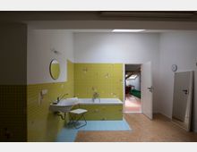 Ein Badezimmer mit gelb gefliester Wand und blauen Bodenfliesen, ausgestattet mit einem Waschbecken, einer Badewanne und einem runden Wandspiegel. Im Hintergrund ist eine offene Tür zu einem angrenzenden Raum mit grünem Teppichboden sichtbar.
