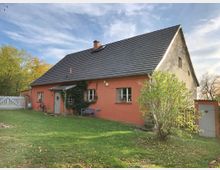 Ein Einfamilienhaus mit oranger Fassade und grauem Satteldach, umgeben von einer Wiese und einigen Bäumen. Vor dem Haus stehen ein Fahrrad, eine blaue Sitzbank und Sträucher, während im Hintergrund ein Holzzaun und ein Schuppen sichtbar sind.