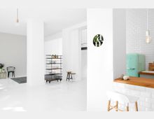 Heller, minimalistischer Innenraum mit weißen Wänden und Böden, einer offenen Küche mit mintgrünem Retro-Kühlschrank und einer Arbeitsfläche aus Holz. Im Raum befinden sich ein metallisches Regal mit Pflanzen, Hocker, ein kleiner Tisch mit Blumen und eine moderne Pendelleuchte.