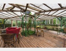 Überdachte Terrasse mit Holzboden, Metall- und Holzkonstruktion, transparentem Wellblechdach und grünen Pflanzen im Inneren. Mehrere Gartenstühle und Tische stehen verstreut, und im Hintergrund sind Fenster sowie eine grüne, bewaldete Umgebung sichtbar.