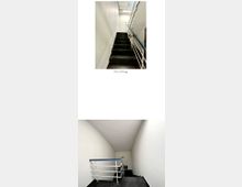 Ein schlichtes Treppenhaus mit weiß gestrichenen Wänden und dunklen, matten Stufen. Ein Metallgeländer mit blauer Handlaufabdeckung verläuft an beiden Seiten, und am oberen Ende ist ein Notausgangsschild sichtbar.