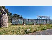 Steinmauer mit einem bemalten Betonzaun im Hintergrund, der eine Landschaft mit Bäumen darstellt; daneben eine Steinstruktur mit einer Skulptur eines Vogels darauf. Im oberen Bereich ist ein Werbeschild für „Tiefbau- und Schüttguthandel“ sichtbar.