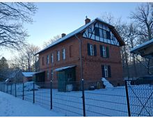 Backsteingebäude mit Fachwerkelementen im Dachgeschoss, umgeben von schneebedecktem Boden und Bäumen. Vor dem Haus befindet sich ein metallener Zaun, und im Hintergrund sind weitere Gebäude und winterliche Waldkulisse zu sehen.