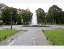 Ein öffentlicher Park mit einem zentralen Springbrunnen, umgeben von Kopfsteinpflasterwegen und gepflegtem Rasen. Im Hintergrund befinden sich große Bäume, eine halbrunde Pergola aus Stein und mehrstöckige Gebäude.