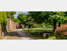 Ein gepflasterter Weg führt entlang einer historischen Steinmauer durch einen parkähnlichen Bereich mit grünen Bäumen und Sträuchern. Im Vordergrund stehen Bänke und eine Laterne, im Hintergrund sind weitere Bäume und Wiesen zu sehen.