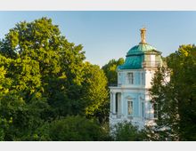 Barockes Gebäude mit blauer Fassade, weißen Säulen und einem kupfergrünen Kuppeldach, das von einer goldenen Skulptur gekrönt ist, umgeben von dichtem, grünem Laub in einem parkähnlichen Umfeld bei klarem Himmel.