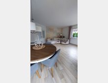 Wohnzimmer mit einem hellen Holzboden, einem runden Holztisch mit grauen Polsterstühlen im Vordergrund und einer gemütlichen Sofaecke mit heller Wandgestaltung im Hintergrund. Rechts befindet sich ein Fenster mit rosa Vorhängen, das Tageslicht hereinlässt.