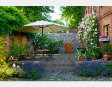 Gepflegter Gartenhof mit einer kleinen Terrasse aus Backstein, auf der ein Holztisch mit Stühlen und ein großer Sonnenschirm stehen. Umgeben von blühenden Rosen, Topfpflanzen und Lavendel, grenzt der Hof an eine Natursteinmauer und ein Backsteinhaus mit gelben Fenstern.