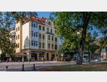 Wohngebäude im Altbaustil mit gelber Fassade, mehreren Balkonen und einem Restaurant im Erdgeschoss, umgeben von Bäumen und einem Gehweg mit Fußgängern. Im Vordergrund befinden sich eine Bushaltestelle, eine Laterne und einige Poller entlang der Straße.