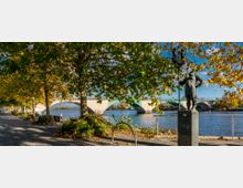 Uferpromenade mit gepflastertem Gehweg, schattenspendenden Bäumen und einer Skulptur aus Metall auf einem Sockel. Im Hintergrund fließt ein breiter Fluss, über den eine steinerne Brücke mit mehreren Bögen führt.