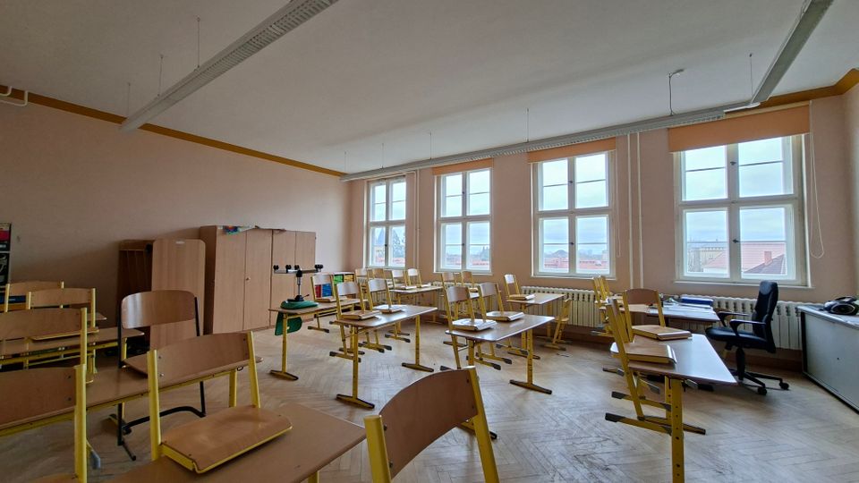 Leeres Klassenzimmer mit hellen Wänden, mehreren Reihen von Schülertischen und hochgestellten Stühlen auf Parkettboden. Große, mehrflügelige Fenster entlang der rechten Wand lassen viel Tageslicht herein; an der Rückwand stehen Schränke und seitlich befindet sich ein Lehrerpult.