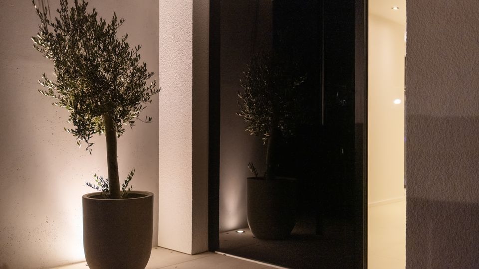 Moderner Hauseingang mit großer dunkler Glasfront und seitlicher Öffnung zu einem hell beleuchteten Innenraum. Vor der Wand steht ein großer Pflanzkübel mit einem kleinen Baum, der von Bodenleuchten angestrahlt wird.
