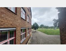 Backsteinfassade eines Gebäudes mit roten Fenstereinfassungen, daneben ein gepflasterter Weg, der an einer Grünfläche mit vereinzelten Bäumen und geparkten Autos vorbeiführt. Im Hintergrund sind weitere Bäume und ein bewölkter Himmel zu sehen.