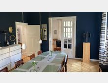 Esszimmer in einem klassischen Interieur mit dunkelblauen Wänden, einem großen Holztisch mit Tischsets und Pendelleuchten darüber. Im Hintergrund eine weiße Tür mit Glaseinsatz, die in einen angrenzenden Raum mit Sesseln führt, flankiert von dekorativen Elementen wie einem goldenen Spiegel und einer Holzskulptur auf einem Sockel.