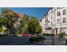 Straßenkreuzung in einem Wohnviertel mit historischen Mehrfamilienhäusern, verziert durch Balkone und rote Ziegeldächer. Im Vordergrund befinden sich ein begrünter Mittelstreifen, parkende Autos und Fußgänger, umgeben von Bäumen und Geschäftseingängen.