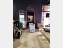 Raum mit Holzboden, dunklen Wänden und einer Glas-/Holztür, die zu einem kleineren Raum führt. Im Vordergrund stehen Möbelstücke wie Barhocker, ein Sessel, sowie ein technisches Gerät und eine Transportkiste.