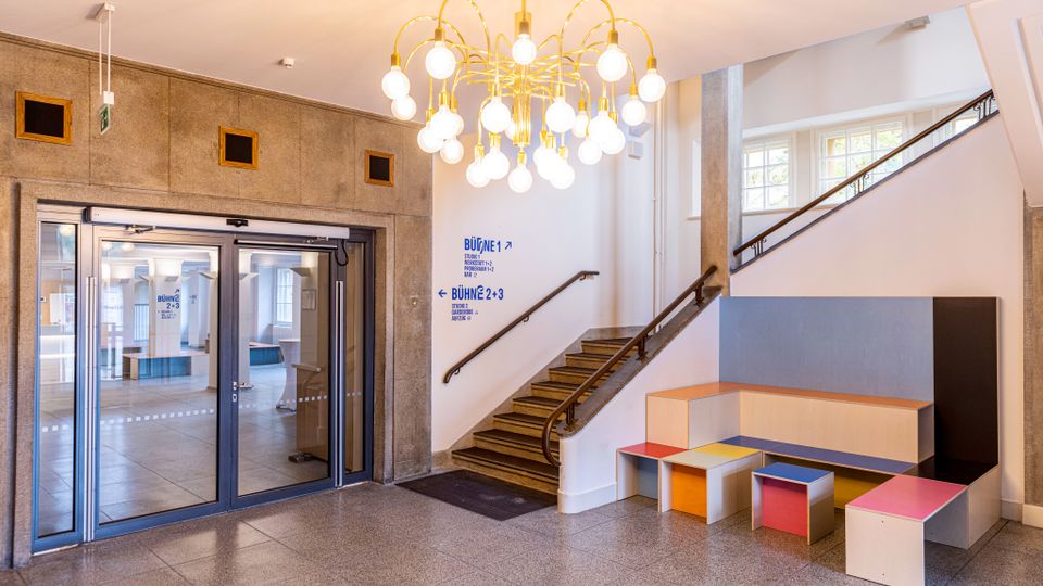 Heller Eingangsbereich mit Glastüren, steinerner Wandverkleidung und einem großen Kronleuchter mit vielen Kugelleuchten. Rechts führt eine Treppe mit Geländer nach oben; daneben stehen modulare Sitzbänke und kleine Hocker in verschiedenen Farben.