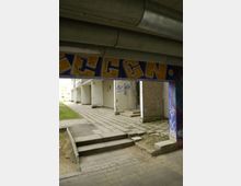 Blick auf den Eingang eines Wohnblocks mit einfacher Betonarchitektur, grauen Fassaden und einer überdachten Passage. Der Boden besteht aus Gehwegplatten, und an der Wand ist Graffiti sichtbar.