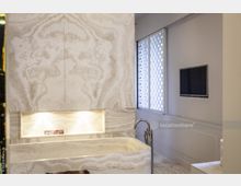 Modernes Badezimmer mit Wänden und einer freistehenden Badewanne aus hellbeigefarbenem Marmor mit natürlicher Maserung, einer eingelassenen, beleuchteten Nische, einem Wandfernseher und großen Fenstern mit gemusterten Sichtschutzpaneelen.