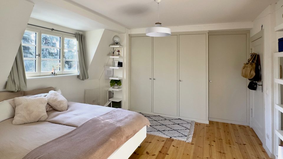 Helles Schlafzimmer mit Holzdielenboden, weißem Bett und großen Fenstern mit Vorhängen an der linken Seite. Gegenüber stehen eingebaute Kleiderschränke mit mehreren Türen; in der Ecke befindet sich ein schmales Regal und eine Deckenlampe hängt mittig.