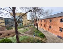 Innenhof mit einem gepflasterten Weg, umgeben von Grünflächen, Bäumen und Sträuchern. Im Hintergrund sind ein modernes Gebäude mit Glasfassade und ein Backsteingebäude mit Satteldach und rechteckigen Fenstern zu sehen.