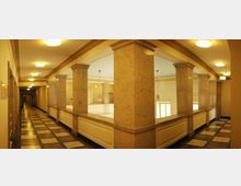 Heller Innenraum eines historischen Gebäudes mit breiten Marmorsäulen, einem Marmorboden mit schwarz-weißen Schachbrettmuster und einer umlaufenden Balustrade. An der Decke sind runde Leuchten und eine hellblaue Färbung erkennbar, die Fenster lassen Tageslicht herein.